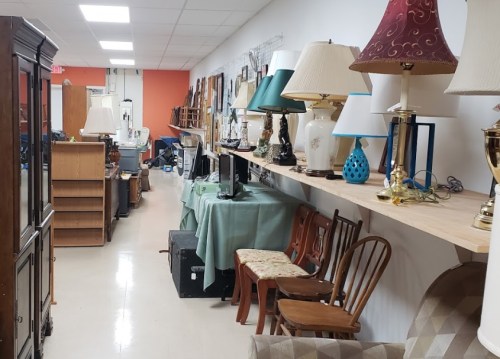 A narrow aisle in a thrift store lined with furniture, lamps, and various home decor items on display.