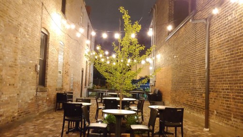 A cozy outdoor dining area with string lights, tables, and a small tree, set between brick walls at night.
