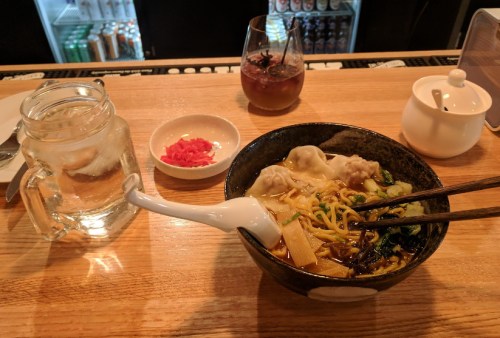 A bowl of noodles with dumplings, chopsticks, a glass of water, and a small dish of pickled ginger on a wooden table.