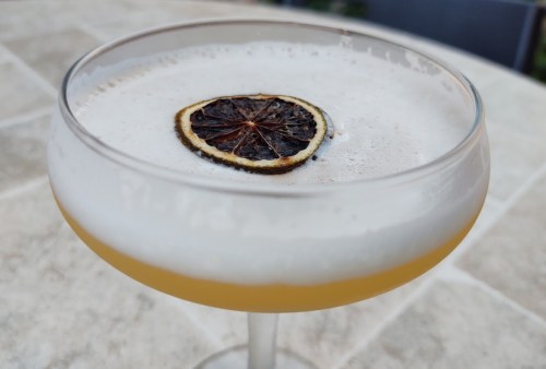 A cocktail in a coupe glass topped with a dried lemon slice, resting on a textured surface.