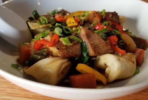 A bowl of dumplings topped with beef, colorful peppers, green onions, and a savory sauce.