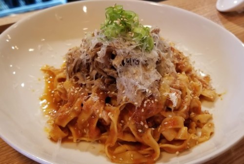 A plate of pasta topped with shredded meat, green onions, sesame seeds, and grated cheese in a rich sauce.