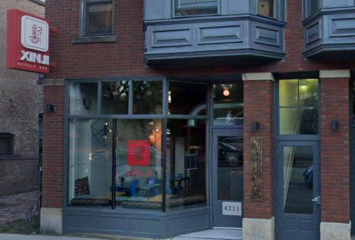 Exterior of XINJ Noodle Bar, featuring a brick facade and large windows displaying the restaurant's logo and interior.