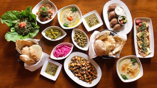 A spread of Middle Eastern dishes including hummus, falafel, olives, pickles, and fresh vegetables on a wooden table.