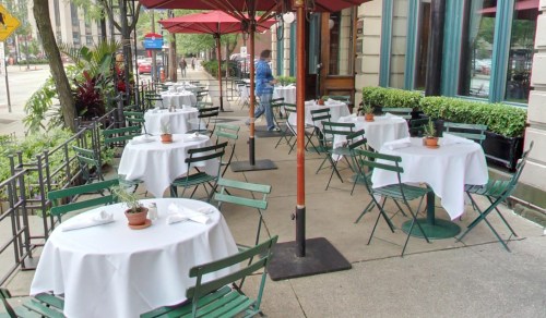 Outdoor dining area with green chairs and white tablecloths, shaded by red umbrellas, in a city setting.