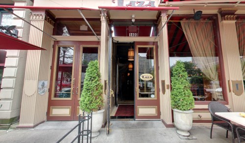 Entrance of a restaurant with double doors, potted plants, and outdoor seating under red awnings.