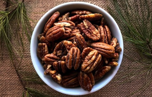A white bowl filled with pecans, surrounded by pine needles on a textured burlap background.