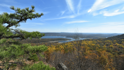 A scenic view of a lake surrounded by autumn foliage and rolling hills under a clear blue sky.