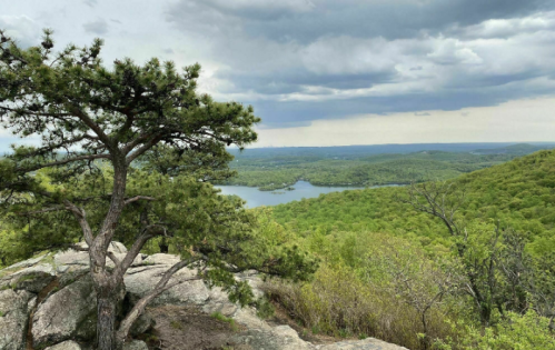 A scenic view from a rocky overlook, featuring a lake surrounded by lush green hills under a cloudy sky.
