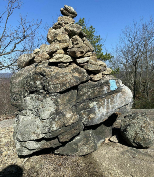 A stacked rock formation on a rocky surface, surrounded by sparse trees and a clear blue sky.