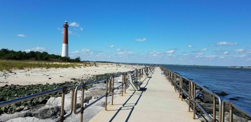A long pier extends towards a lighthouse by the shore, with sandy beach and water under a blue sky.