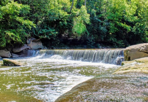 A serene waterfall cascades over rocks into a calm river, surrounded by lush green trees and foliage.