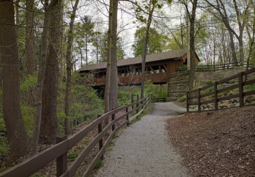 A gravel path leads to a wooden covered bridge surrounded by trees and greenery.