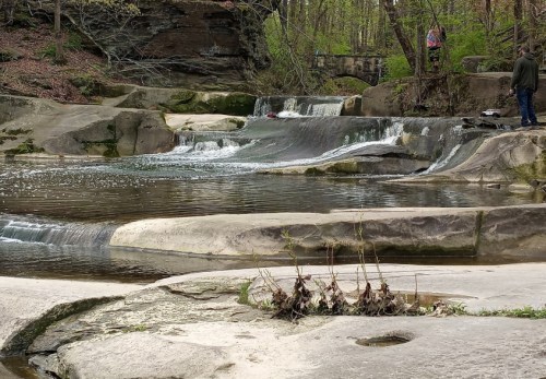 A serene waterfall cascades over smooth rocks, surrounded by trees, with two people in the background enjoying the scenery.