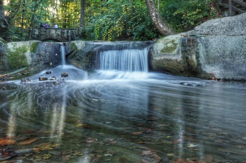 A serene waterfall cascades over rocks into a clear pool, surrounded by lush green trees and autumn leaves.