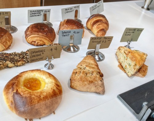 A display of assorted pastries, including croissants, scones, and a danish, with price tags on a wooden table.
