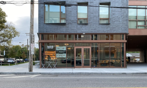 A modern storefront with large windows, featuring a sign that says "NOW OPEN" and outdoor seating.