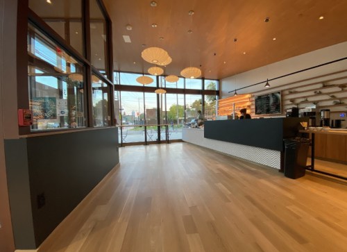 A modern café interior with wooden floors, large windows, and pendant lights, featuring a service counter and seating area.
