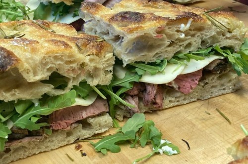 Close-up of a sliced sandwich with layers of roast beef, mozzarella, and arugula on crusty bread.