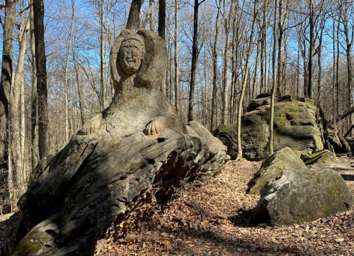 A carved stone figure resembling a seated person, surrounded by trees and rocky terrain in a forest setting.