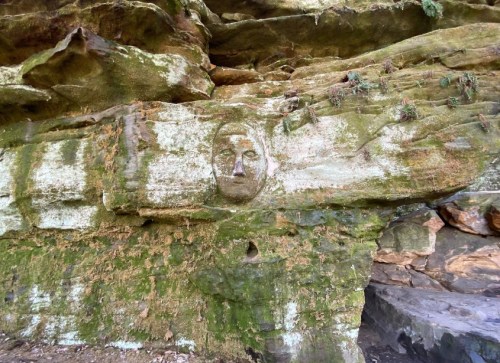 Carved face on a moss-covered rock wall, surrounded by natural stone formations and greenery.