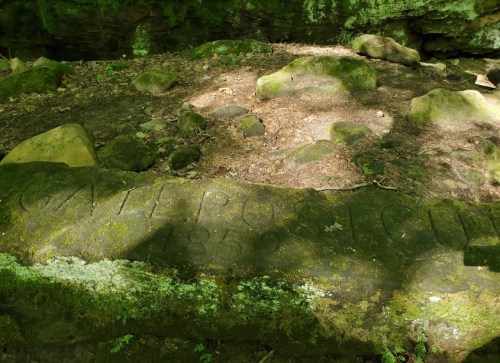 A moss-covered stone with the inscription "GATE POST 1852" surrounded by rocks and forest floor.