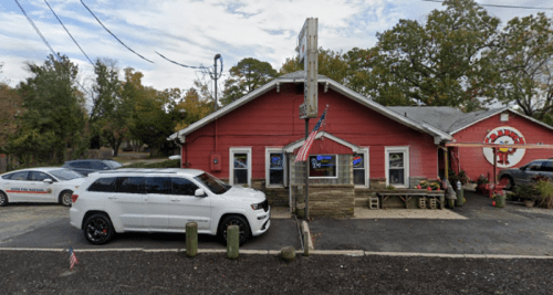 A pink building with a sign, parked white SUV, and American flags outside, surrounded by trees and a gravel lot.
