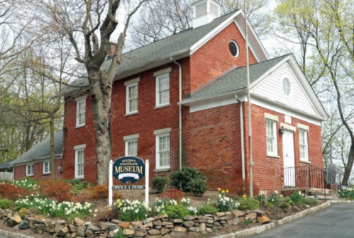 A red brick museum building with white trim, surrounded by blooming flowers and greenery.