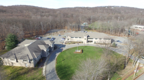 Aerial view of a building complex surrounded by trees and a parking lot, with a grassy area in the foreground.