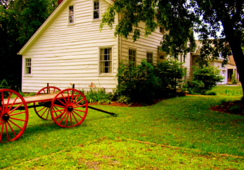 A white house with green surroundings and a red-wheeled cart in the foreground, set in a lush garden.
