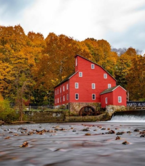 A red mill surrounded by vibrant autumn foliage and a flowing river, creating a picturesque fall scene.