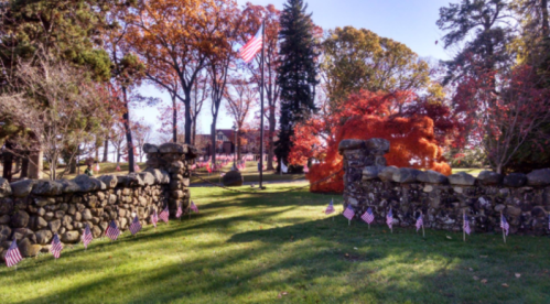 A stone entrance with American flags leads to a park with colorful autumn trees and a flagpole in the background.