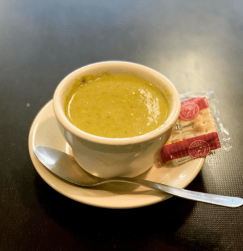 A bowl of green soup on a plate, accompanied by a small packet of crackers and a spoon.