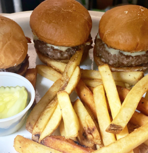 Three mini burgers with cheese on a plate, accompanied by a side of golden French fries and a small cup of pickles.