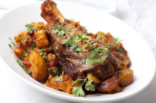 A plate of tender lamb shank served with potatoes and a rich tomato sauce, garnished with fresh herbs.