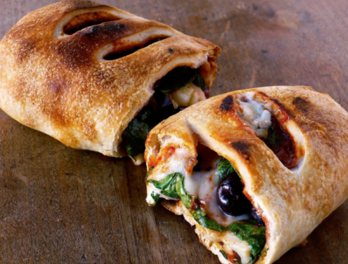 A golden-brown calzone filled with spinach, cheese, and olives, partially sliced open on a wooden surface.
