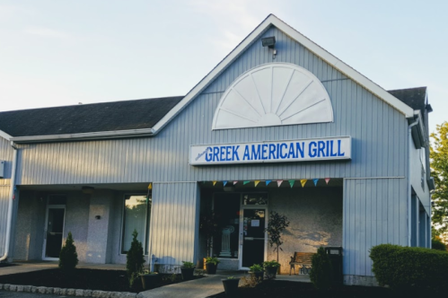 Exterior of Greek American Grill restaurant, featuring a blue sign and decorative plants in front.