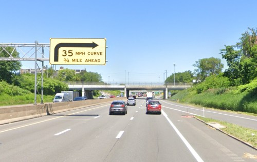A highway scene with a sign indicating a 35 MPH curve ¾ mile ahead, surrounded by greenery and vehicles on the road.