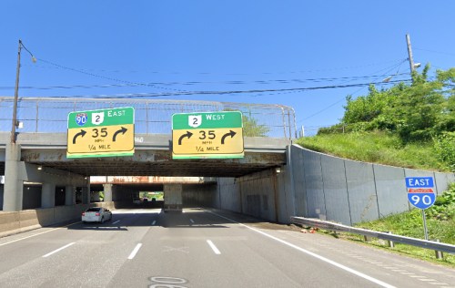 Road signs indicating I-90 East and West directions, with a clear blue sky above and a vehicle approaching.