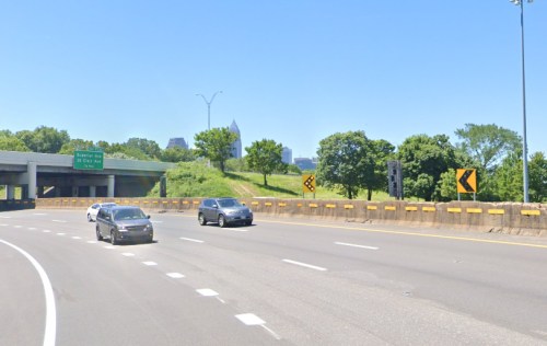 Cars driving on a highway with a clear blue sky and city skyline in the background. Green trees line the roadside.