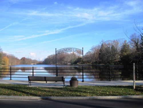 A peaceful view of Brainerd Lake with a sign, bench, and trees reflecting in the water under a blue sky.