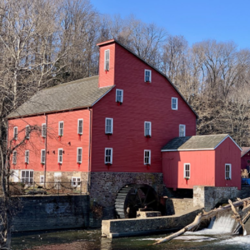 A red historic mill with a water wheel, surrounded by trees and a flowing river on a clear day.