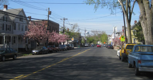A tree-lined street with blooming cherry blossoms, parked cars, and a clear blue sky.