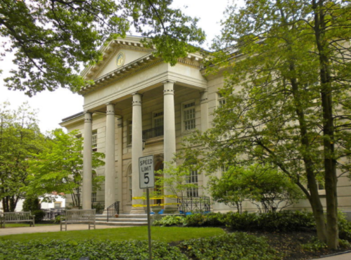 A classic building with columns, surrounded by trees and greenery, featuring a speed limit sign in front.