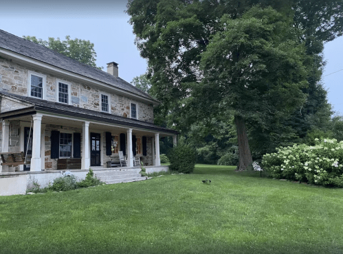 A stone house with a porch, surrounded by lush green grass and trees, under a cloudy sky.