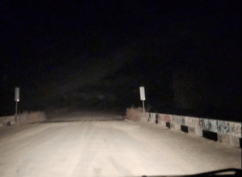 A dark, unlit road leading to a graffiti-covered bridge at night, with faint signs visible on either side.