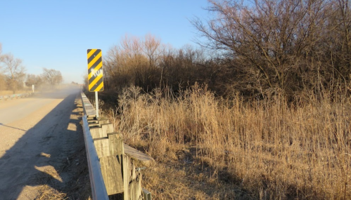 A gravel road with a warning sign beside a fence, surrounded by dry grass and sparse trees under a clear blue sky.