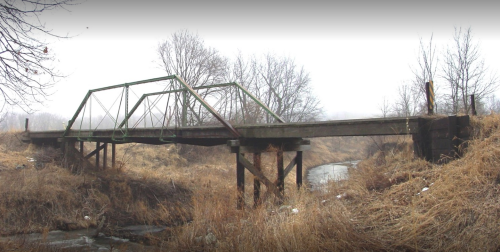 An old, green truss bridge spans a small creek, surrounded by dry grass and bare trees in a foggy landscape.