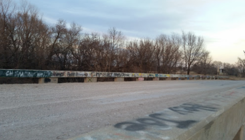 A weathered bridge with graffiti on the railing, surrounded by bare trees and a gravel path.