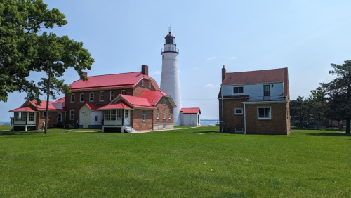 A lighthouse stands tall behind two historic brick buildings with red roofs, surrounded by green grass and trees.
